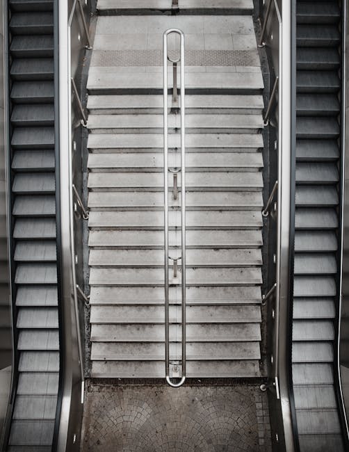 An overhead view of a set of staircases inside a building, showing metal handrails running down the center and concrete steps with a non-slip surface, leading from a lower level to an upper floor. The
