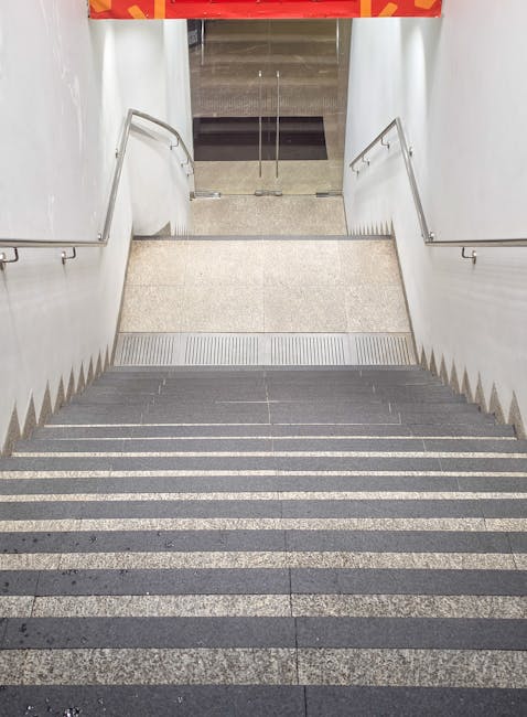 An overhead view of a set of staircases inside a building, showing metal handrails running down the center and concrete steps with a non-slip surface, leading from a lower level to an upper floor. The