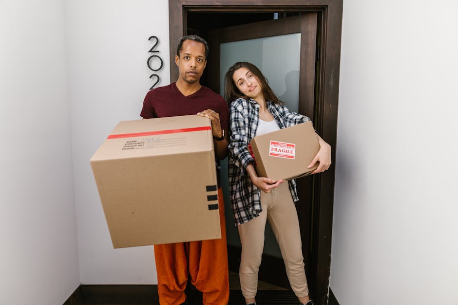 A man and a woman standing side by side indoors near a doorway, each holding cardboard boxes for home relocation. The man is wearing a burgundy t-shirt and orange trousers, holding a large box with black tape on the edges. The woman is dressed in a checkered shirt over a white top and beige trousers, holding a smaller box labeled 'Fragile' with red tape. Both are positioned on a dark floor, with the doorway behind them featuring a dark wood frame and a frosted glass panel. The background wall is plain, and the lighting suggests an interior environment. The scene depicts packing and moving processes typical of professional removals services, such as those provided by Clapham Junction Removals, highlighting furniture transport and the careful handling of boxed items during house relocations.